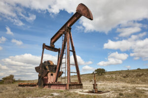 Old oil pump jack standing idle in a rural field under a partly cloudy sky.